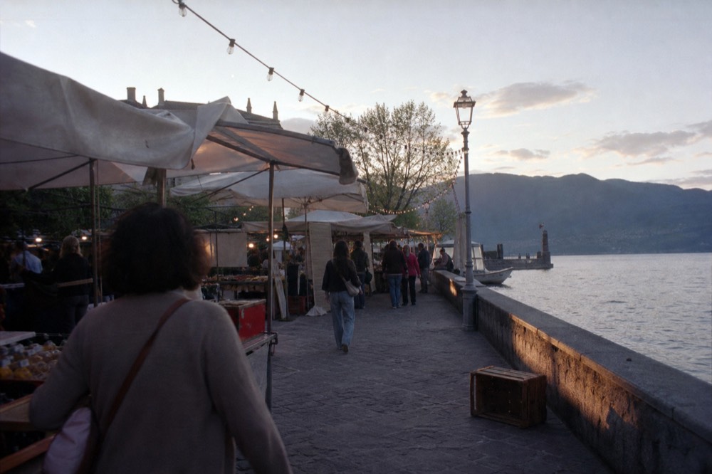 A lakeside festival square with market stalls under strings of lights, no text, no logos, no people