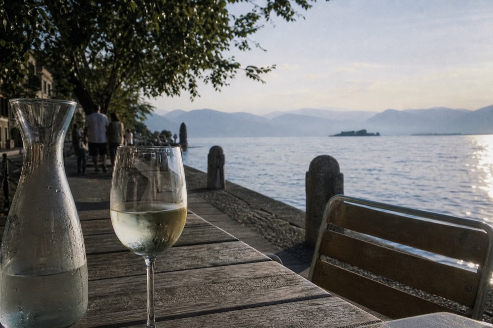 A glass of pale straw-colored Lugana on a lakeside table beside a water carafe, no text, no logos, no people